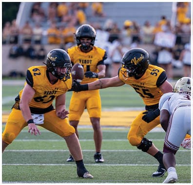 Four football players in yellow uniforms and one player in a white uniform are positioned on a field during a game with a crowd and cheerleaders in the background. Descripci n generada con IA