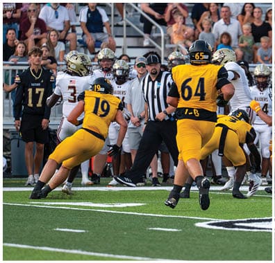 Players in yellow and white football uniforms are actively engaged in a play on a green field with spectators and officials watching in the background. Descripci n generada con IA
