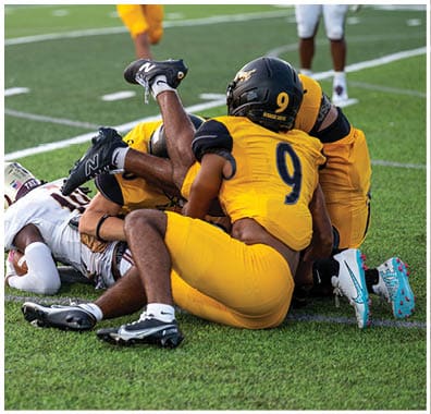 Two people in football uniforms are on the ground during a game on a green field with white yard lines. Descripci n generada con IA