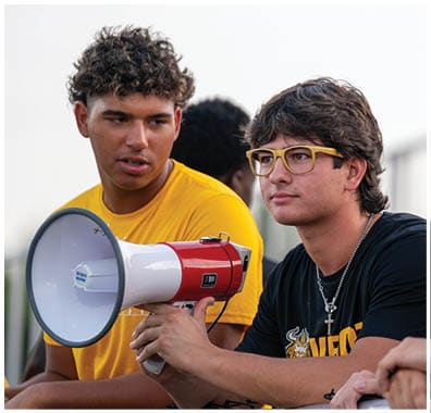 Two people are engaged in conversation outdoors, one wearing a yellow shirt and the other wearing yellow-framed glasses. Descripci n generada con IA