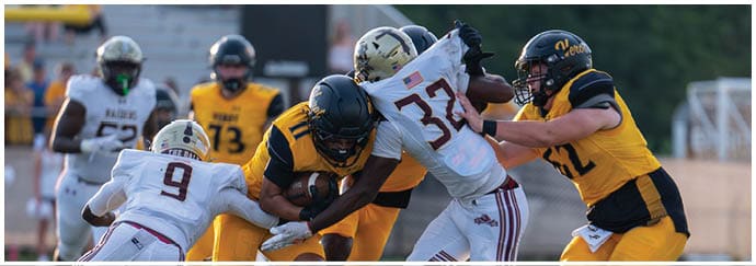 People wearing football uniforms are engaged in a tackle during a game on a grassy field. Descripci n generada con IA
