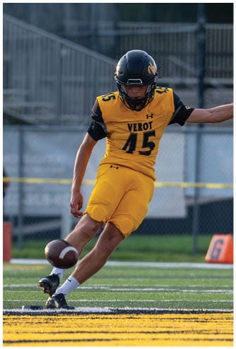 A person in a yellow and black football uniform is kicking a football on a field with other players and spectators in the background. Descripci n generada con IA