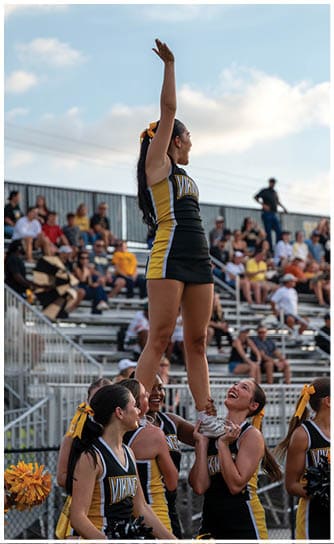 A person in a cheerleading uniform with a bow in their hair is raising one arm and appears to be shouting or cheering at an outdoor event with a crowd in the background. Descripci n generada con IA