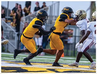 Two people in yellow football uniforms are actively engaged in a play against two people in white football uniforms on a field with spectators in the background. Descripci n generada con IA