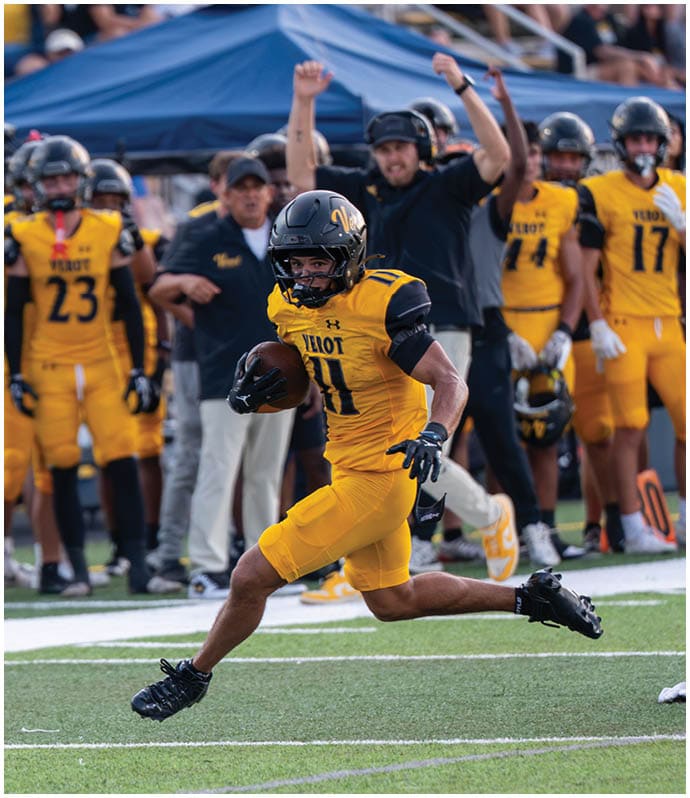 A person in a yellow football uniform is running on a field while holding a football, with teammates and coaches in similar uniforms and attire cheering in the background. Descripci n generada con IA