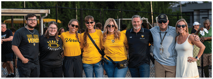 A group of eight adults wearing casual and sports-themed clothing stand closely together outdoors, smiling in front of a fence with trees and a building in the background. Descripci n generada con IA