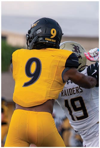 Two people in football uniforms are engaged in a close physical play, with one wearing a yellow and black uniform and the other in a white and maroon uniform. Descripci n generada con IA