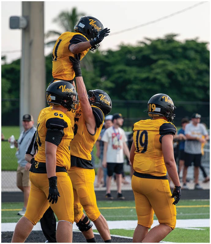 People wearing yellow and black football uniforms celebrate on a field with one person being lifted into the air by teammates. Descripci n generada con IA