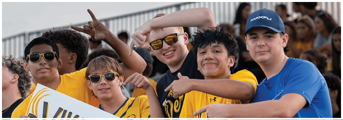 A group of smiling people wearing casual clothing and sunglasses are posing closely together outdoors with a crowd in the background. Descripci n generada con IA