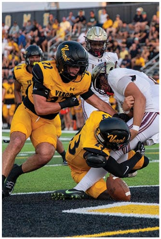 Two people in yellow football uniforms tackle a person in a white uniform near the end zone on a football field while a crowd watches from the stands. Descripci n generada con IA