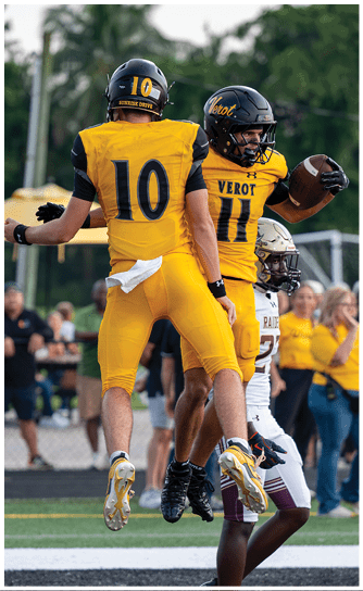 Two adults in yellow football uniforms with black helmets are on a field, one holding a football and the other standing nearby. Descripci n generada con IA