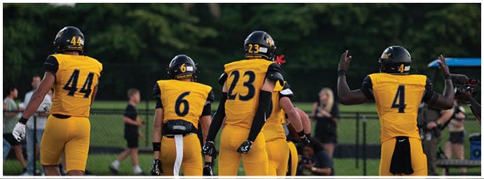 Four people wearing yellow and black football uniforms with helmets are standing on a field with trees in the background. Descripci n generada con IA