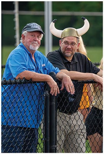 Two adults are leaning on a chain-link fence outdoors, one wearing a blue shirt and cap, and the other wearing a black shirt and a Viking helmet with horns. Descripci n generada con IA