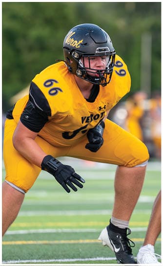 A person in a yellow and black football uniform with the number 66 is crouched in a ready position on a football field facing an opponent. Descripci n generada con IA