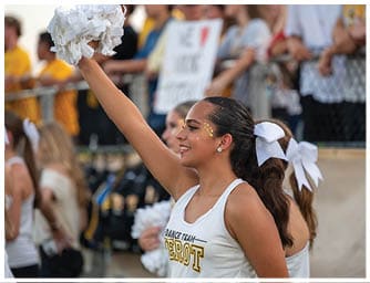 A person with a ponytail and a white bow is smiling and raising a pom-pom while wearing a white tank top with “Dance Team" written on it, with other similarly dressed people and a crowd in the background. Descripci n generada con IA