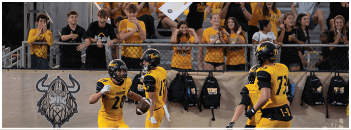 Four people in yellow and black football uniforms are on a field near a wall with a Viking logo while a crowd of people in yellow shirts watches from behind a fence. Descripci n generada con IA
