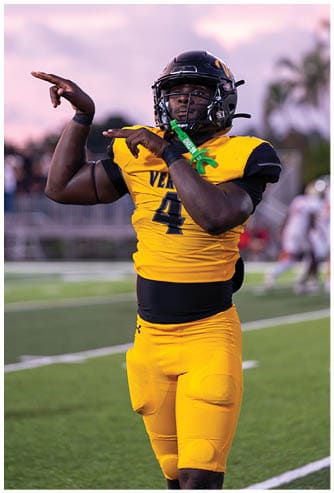 A person wearing a yellow and black football uniform is preparing to throw a football on a field with blurred players and palm trees in the background. Descripci n generada con IA