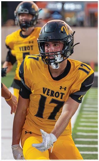 A person in a yellow football uniform with the number 17 and the word Verot is sitting on the sideline reaching out while another person in casual clothing stands nearby. Descripci n generada con IA
