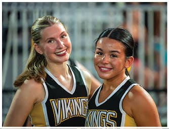 Two adults wearing matching sports jerseys with the word Vikings smile and pose closely together in an outdoor setting. Descripci n generada con IA
