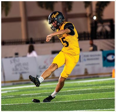 A person in a yellow and black football uniform is kicking a football on a green field during a game. Descripci n generada con IA