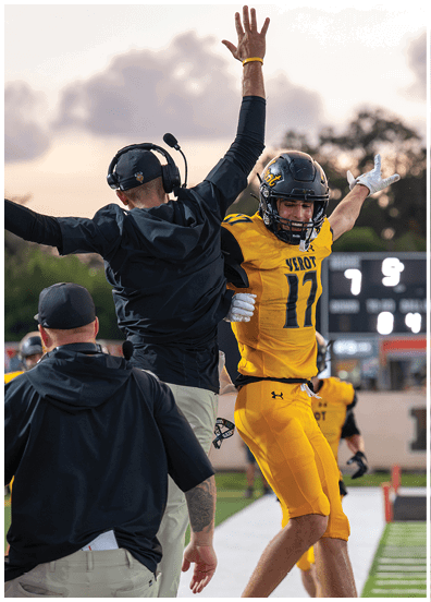 A person in a yellow football uniform celebrates with arms raised while two coaches in black clothing also raise their arms on the sidelines of a football field. Descripci n generada con IA