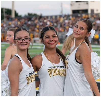 Three people wearing white dance team uniforms with “VERO" printed on them smile and pose together on a sports field with a crowd in the background. Descripci n generada con IA