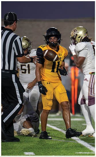 A person in a yellow football uniform catches a football while surrounded by people in white uniforms and a referee on a football field at dusk. Descripci n generada con IA