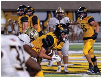 People wearing football helmets and uniforms are on a field with some standing in the background and others closer to the camera. Descripci n generada con IA