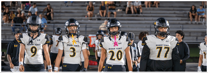 Four people wearing football helmets and Bishop Verot jerseys stand side by side on a football field with empty bleachers and scattered spectators in the background. Descripci n generada con IA