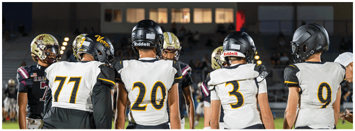Five people wearing football helmets and uniforms stand on a field facing away from the camera with a group of people in the background. Descripci n generada con IA