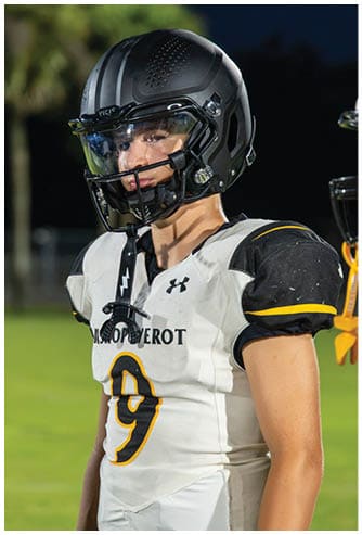 A person wearing a black football helmet and a white football jersey with the number nine stands on a grassy field at night. Descripci n generada con IA
