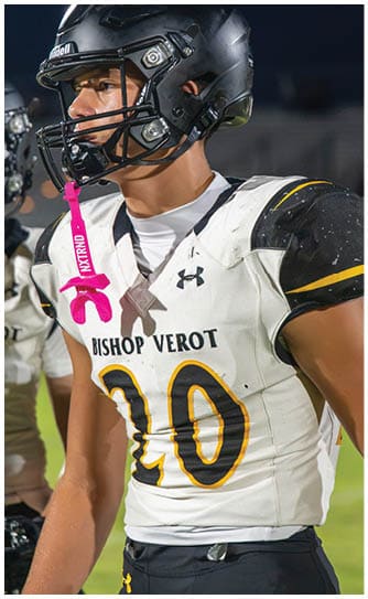 A person wearing a white and black football uniform with the number 20 and the words Bishop Verot is standing on a field with a pink mouthguard hanging from their helmet. Descripci n generada con IA