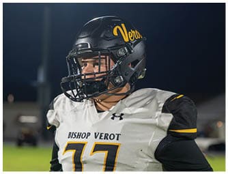 A person wearing a black football helmet and a white Bishop Verot jersey with the number 77 stands on a field at night. Descripci n generada con IA