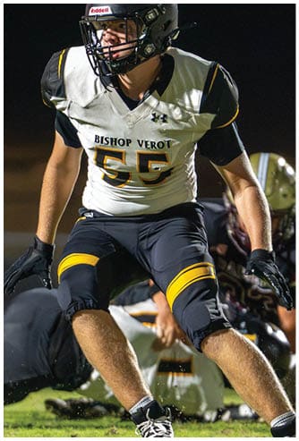 A person wearing a black helmet and a white football uniform with “Bishop Verot" on the jersey is running on a field with another player blurred in the background. Descripci n generada con IA