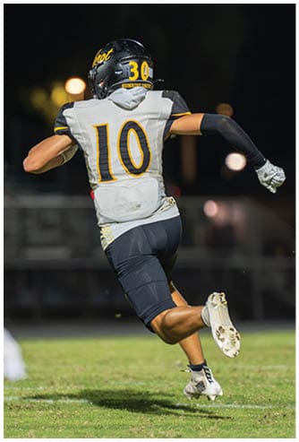 A person wearing a black football helmet and white Bishop Verot jersey with the number 66 stands on a football field with blurred players and bleachers in the background. Descripci n generada con IA