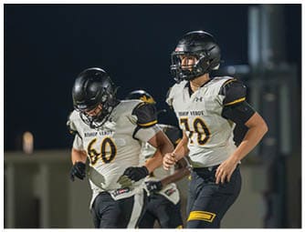 Two people in football uniforms are engaged in a tackle on a field while another player in uniform runs nearby with spectators in the background. Descripci n generada con IA