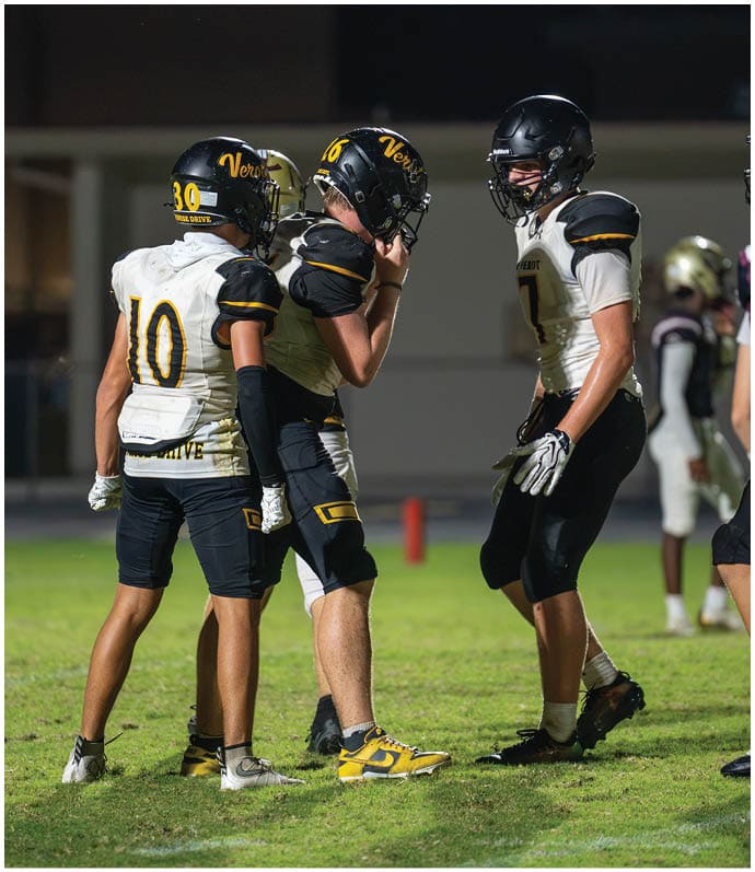 Two people in football uniforms are actively playing on a field at night, with one kicking the ball and the other preparing to block. Descripci n generada con IA