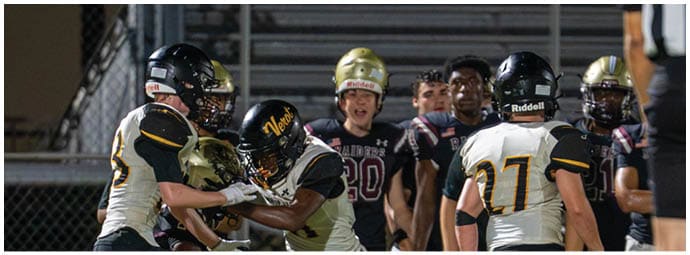People in football uniforms are actively engaged in a play on a field with bleachers and spectators in the background. Descripci n generada con IA