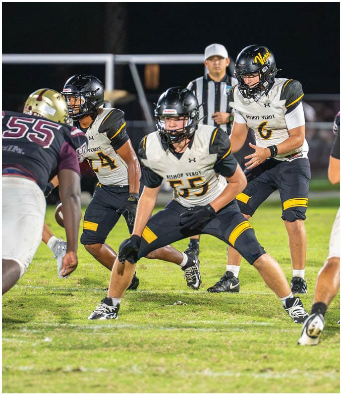 Two people in white sports uniforms with black helmets are running on a football field while two others in green uniforms are nearby. Descripci n generada con IA