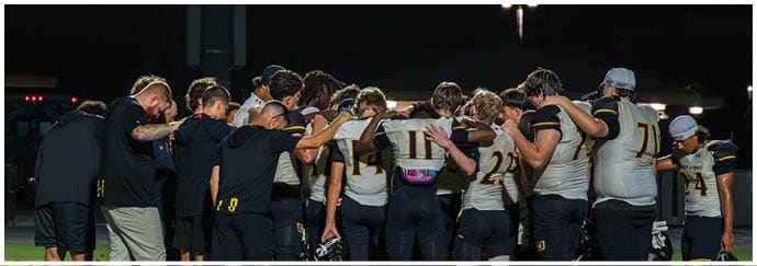 A group of people in football uniforms are kneeling on a field at night while two people stand nearby, one speaking into a microphone. Descripci n generada con IA