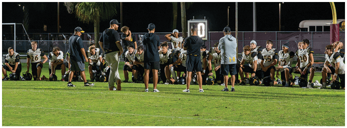 A group of people in sports uniforms and casual clothing are gathered on the sidelines of a football field, some standing and some sitting, with a goalpost and bleachers in the background. Descripci n generada con IA