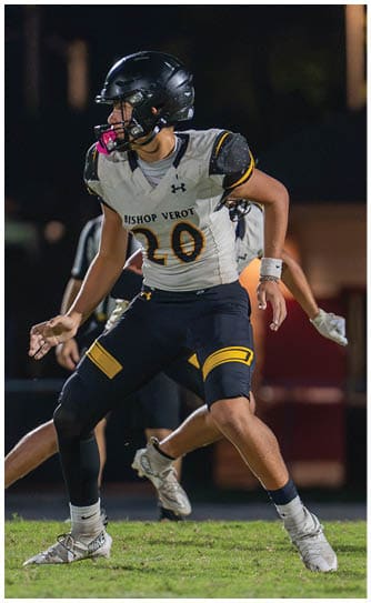 People in football uniforms are actively engaged in a nighttime game on the field with one player in white attempting to block or reach over players in green. Descripci n generada con IA