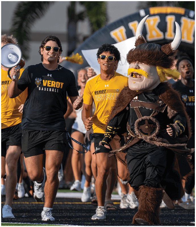Adults wearing sports apparel and sunglasses are running alongside a person in a Viking mascot costume on a sports field. Descripci n generada con IA