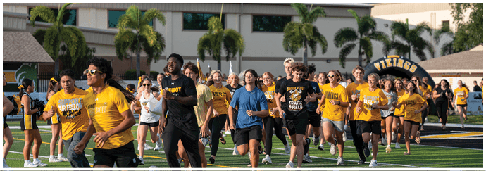 A group of people wearing athletic clothing are running or jogging together on a sports field with palm trees and buildings in the background. Descripci n generada con IA