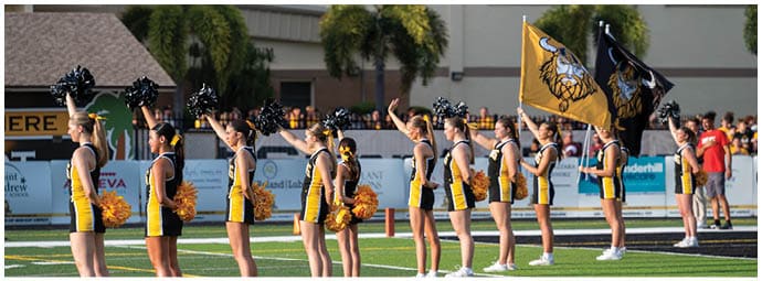 Four people wearing football helmets and white jerseys with “Bishop Verot" written on them stand side by side on a sports field with empty bleachers in the background. Descripci n generada con IA