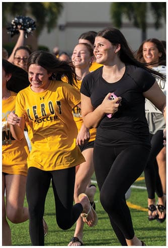 A group of people wearing casual athletic clothing are running and smiling on a grassy field during a sunny day. Descripci n generada con IA