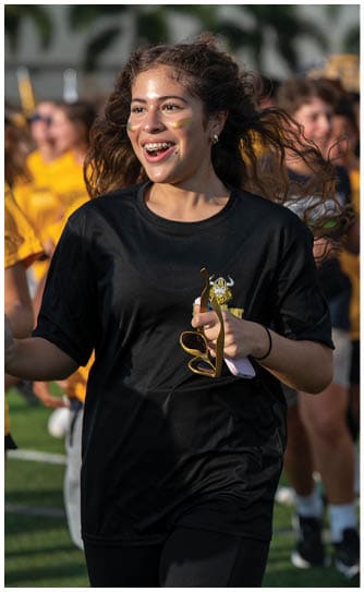 A person with long hair wearing a black shirt and black pants is smiling and holding a small trophy while walking on a field with other people in the background. Descripci n generada con IA