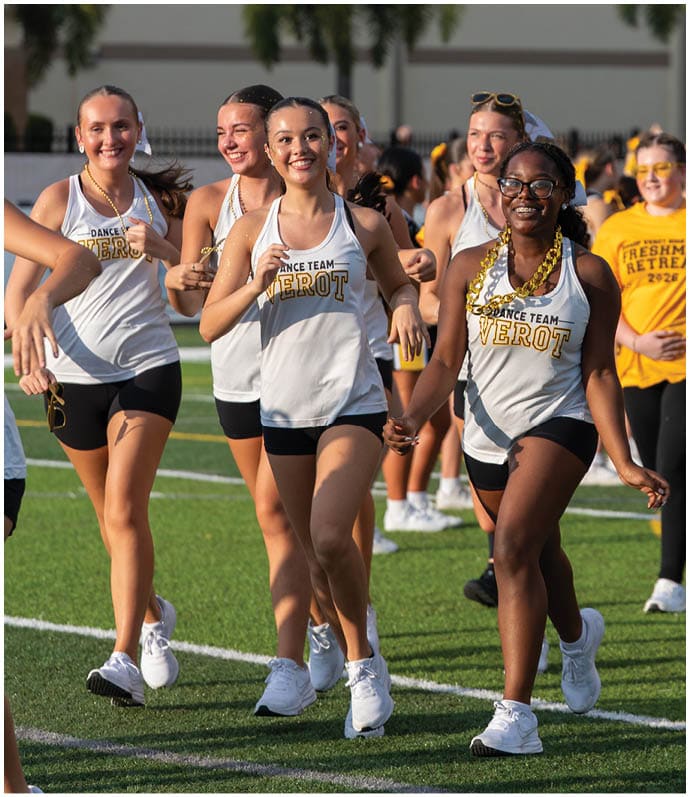 A group of people wearing matching dance team tank tops and shorts are smiling and walking together on a sports field with one person in the background wearing a yellow shirt that says Freshman Retreat. Descripci n generada con IA