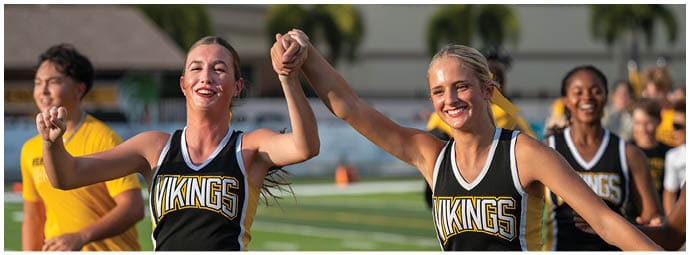 Two adults wearing matching Vikings cheerleading uniforms are smiling and holding hands while performing on a sports field with other people in the background. Descripci n generada con IA