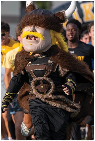 A person wearing a large Viking-style mascot costume with a horned helmet and yellow mustache is running outdoors with a group of people behind them. Descripci n generada con IA